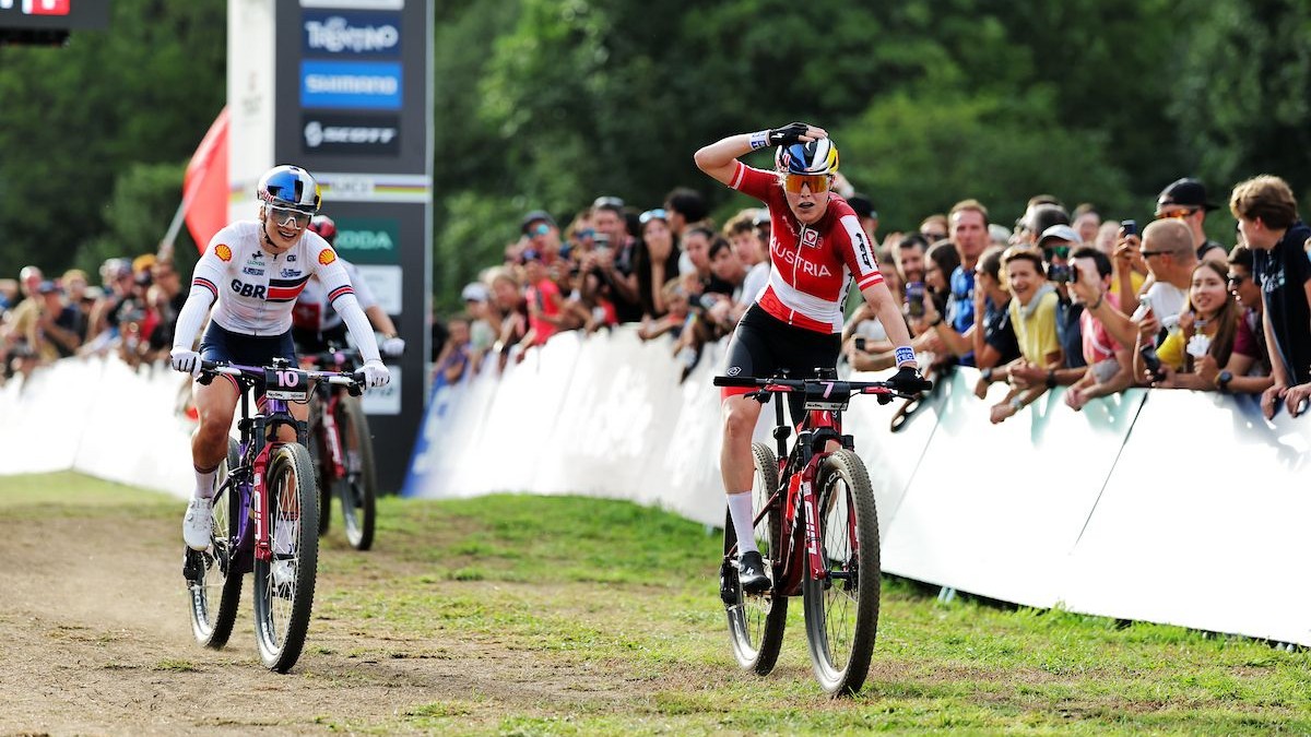 Laura Stigger, com o uniforme vermelho e branco da Áustria, leva a mão ao capacete ao cruzar a linha de chegada à frente de Evie Richards, com o público atrás das grades.